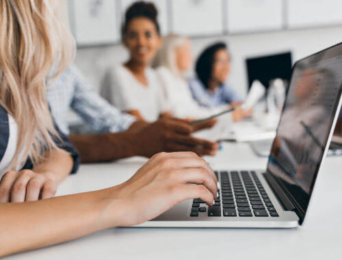In primo piano la mano di una donna che digita qualcosa su un PC portatile in un ufficio. Sullo sfondo altri membri del team
